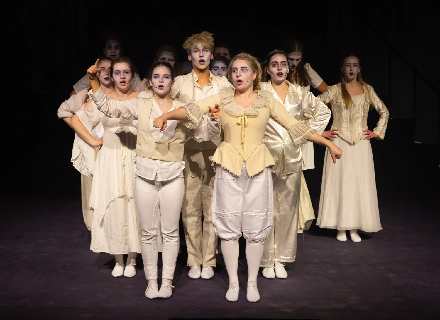 Group of young performers dressed in white, standing on stage in a theatrical production, some with painted faces.