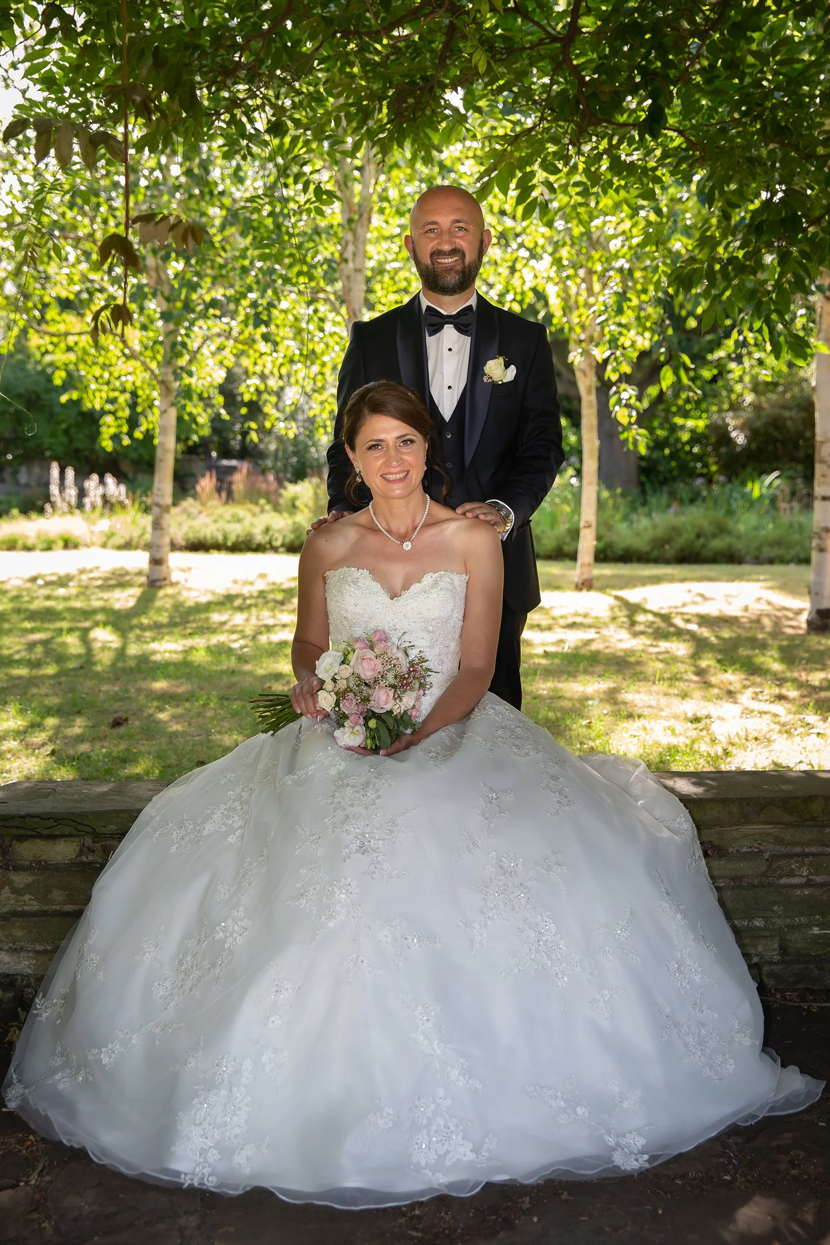A bride and groom pose outdoors for their wedding photo. The bride sits in a white lace wedding gown holding a flower bouquet, and the groom stands behind her in a black tuxedo with a bow tie. They are under a canopy of green trees.