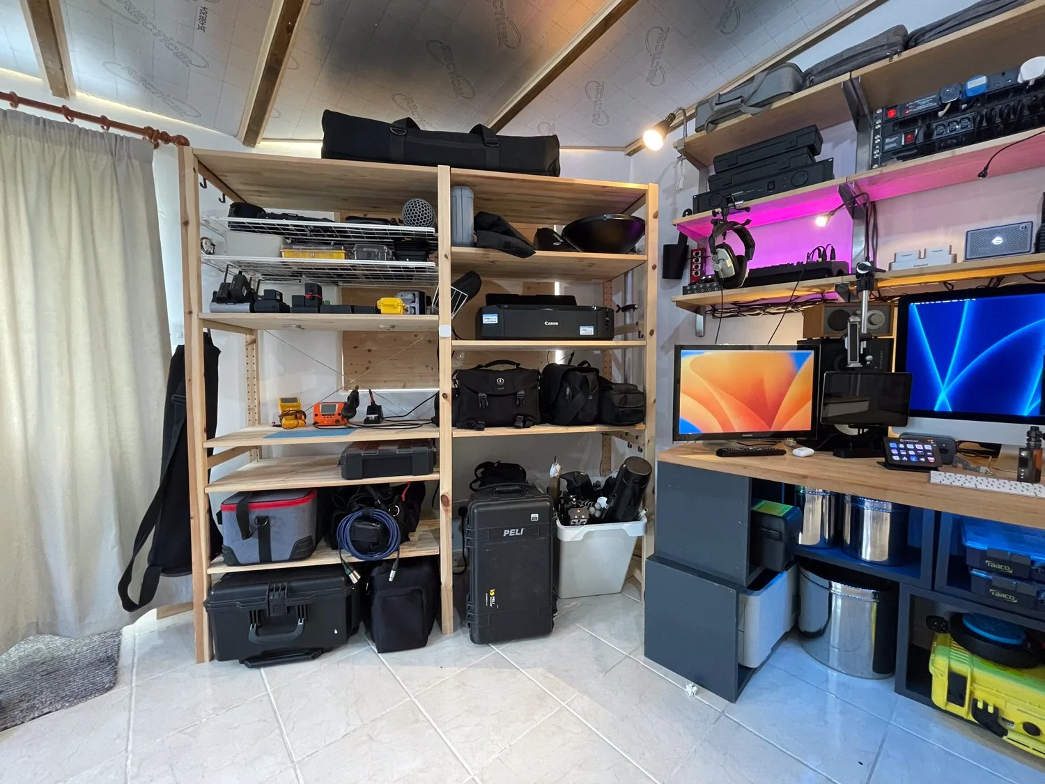 A room with wooden shelves filled with electronic equipment, tools, and bags, and a desk with computers and accessories, with a curtain on the left side.