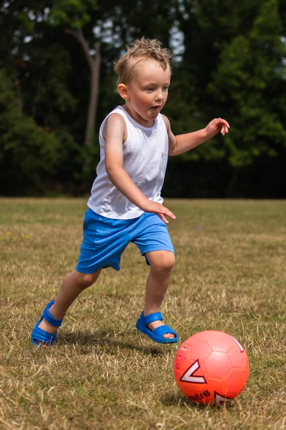 A young boy wearing a white sleeveless shirt, blue shorts, and blue sandals playing soccer outdoors on a grassy field with trees in the background.