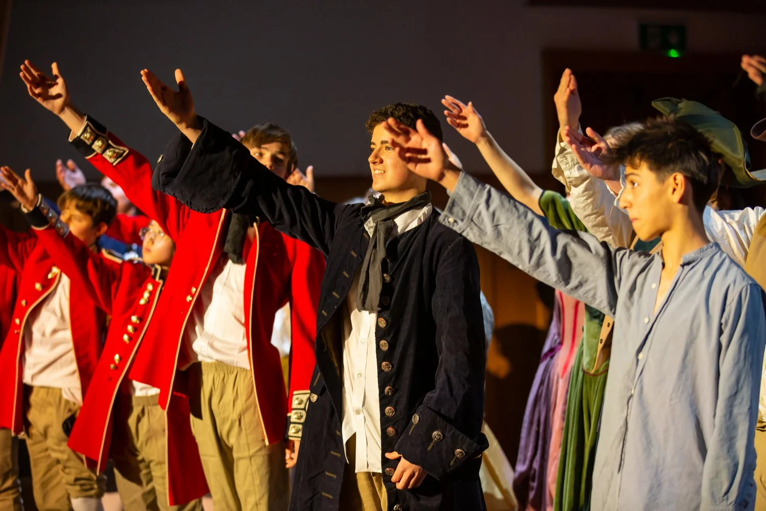 Children in costume on stage performing or participating in a theatrical or historical reenactment, with arms raised.