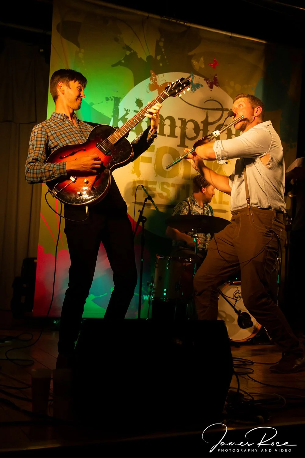 A band performs live on stage, featuring a guitarist and a flutist in the foreground, with a drummer in the background, under colorful stage lighting.