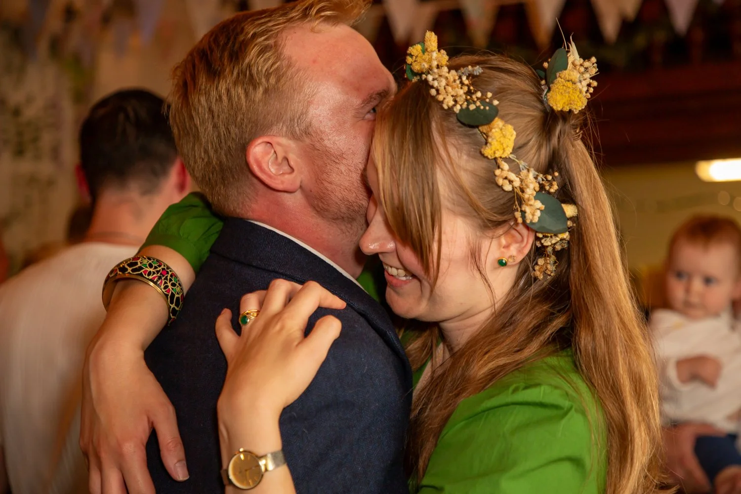A couple dancing closely at a celebration, with one woman wearing a floral crown and bright green top, and a man in a dark suit. In the background, a child and another person are visible.