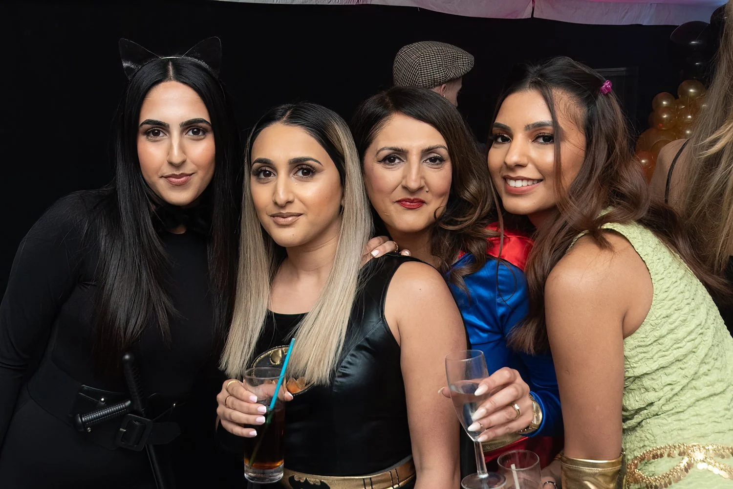 Four women dressed in costumes at a party, holding drinks and smiling at the camera, with balloons and other partygoers in the background.