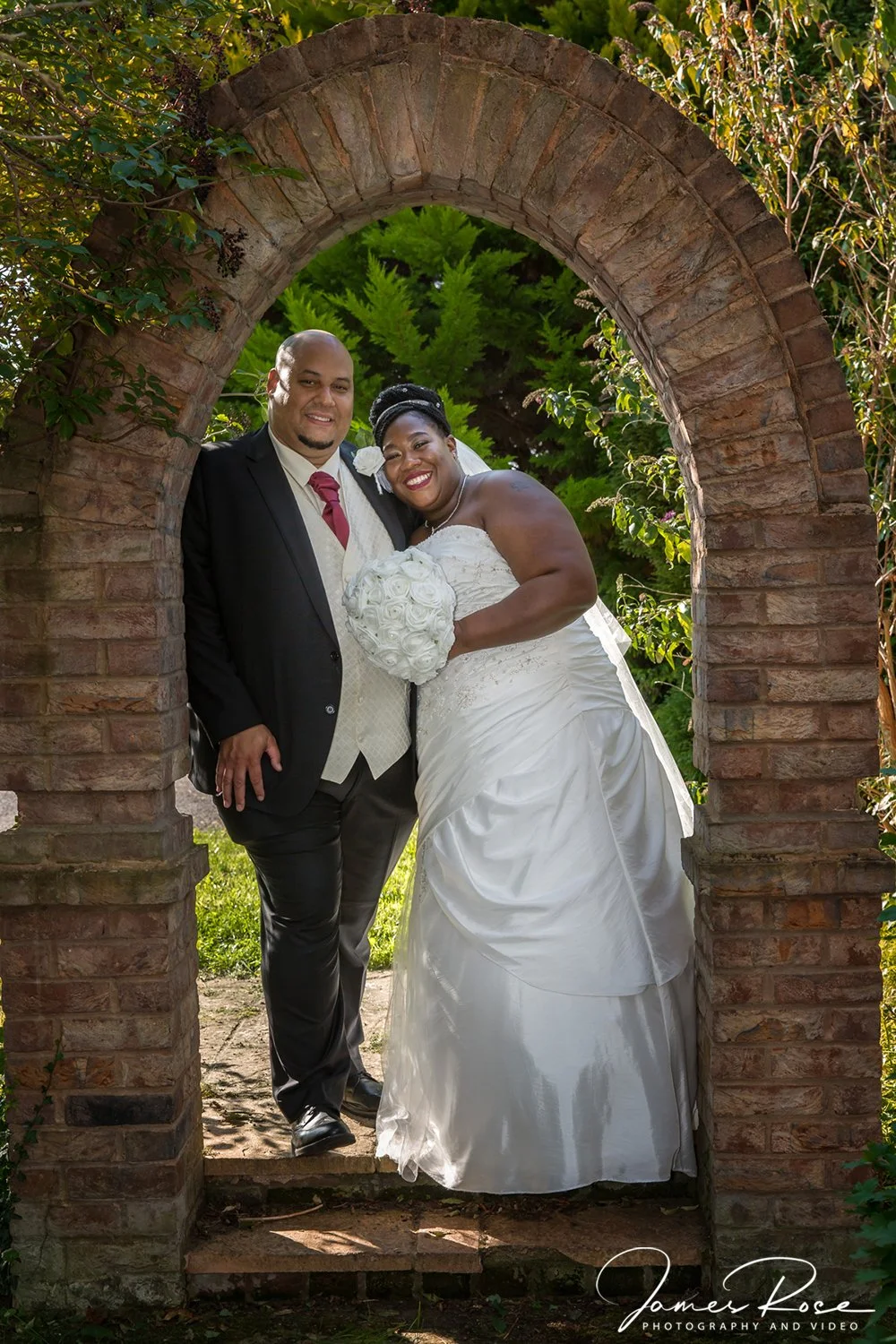 A smiling newlywed couple standing under a brick archway, the bride holding a bouquet of white roses, in a garden with green foliage.