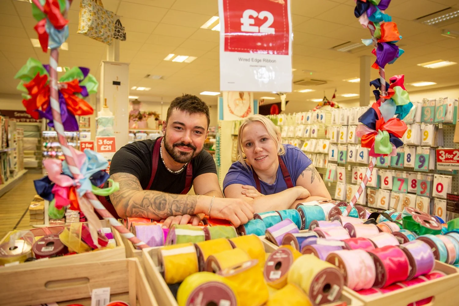 Two store employees, one male and one female, smiling and leaning on a display of colorful ribbons inside a craft store.