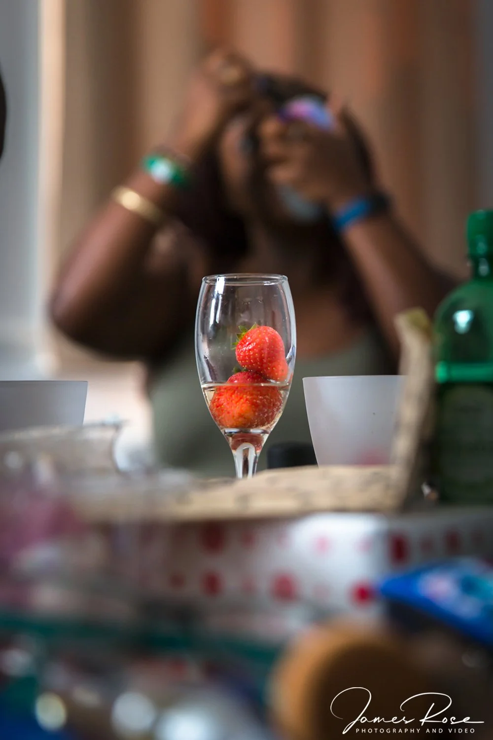 A glass of champagne with strawberries inside, on a table, with a blurred person taking a photo in the background.