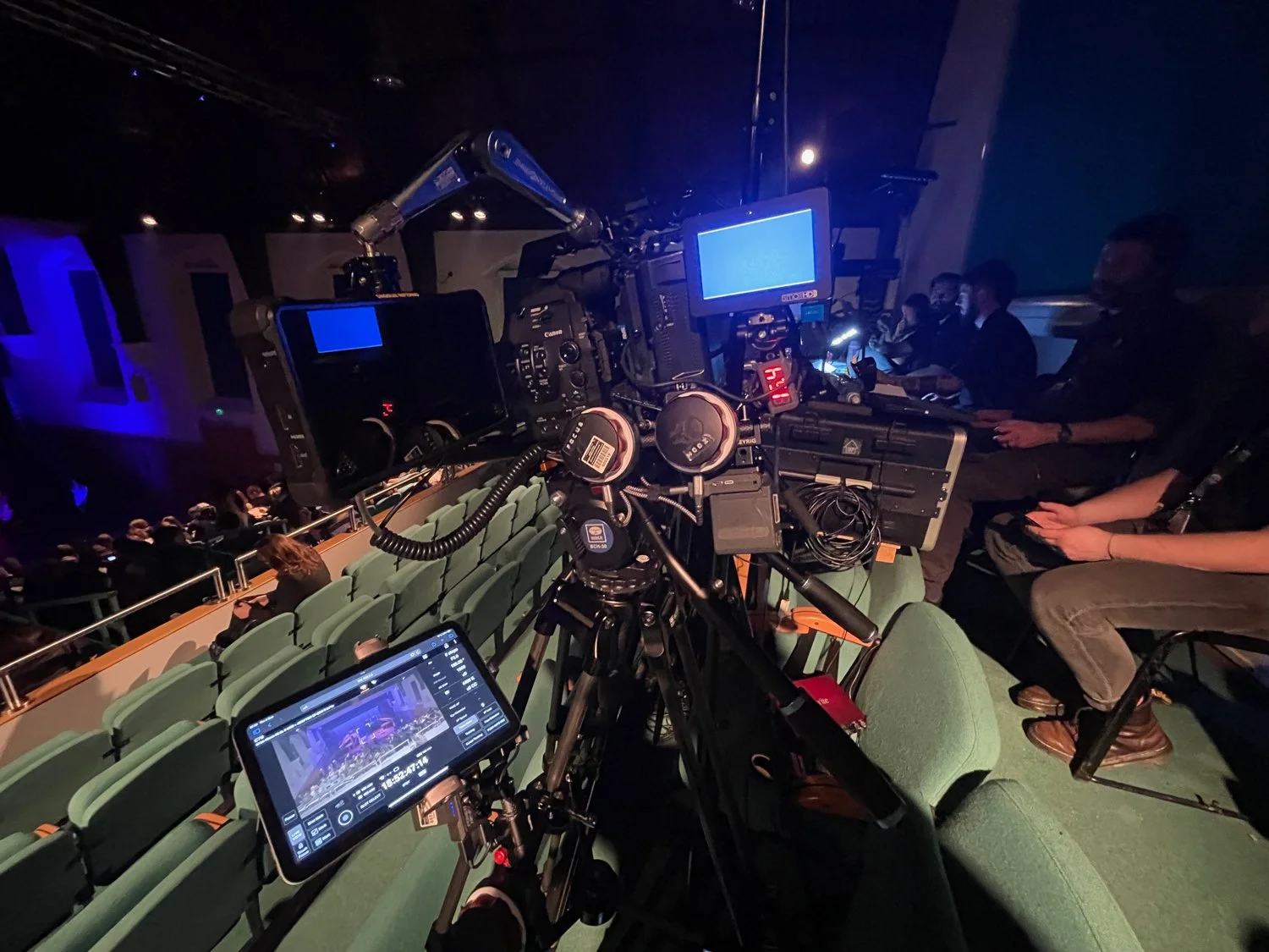View of a professional video camera setup at an indoor event with audience seated in the background, and several crew members working with equipment in the dimly lit environment.