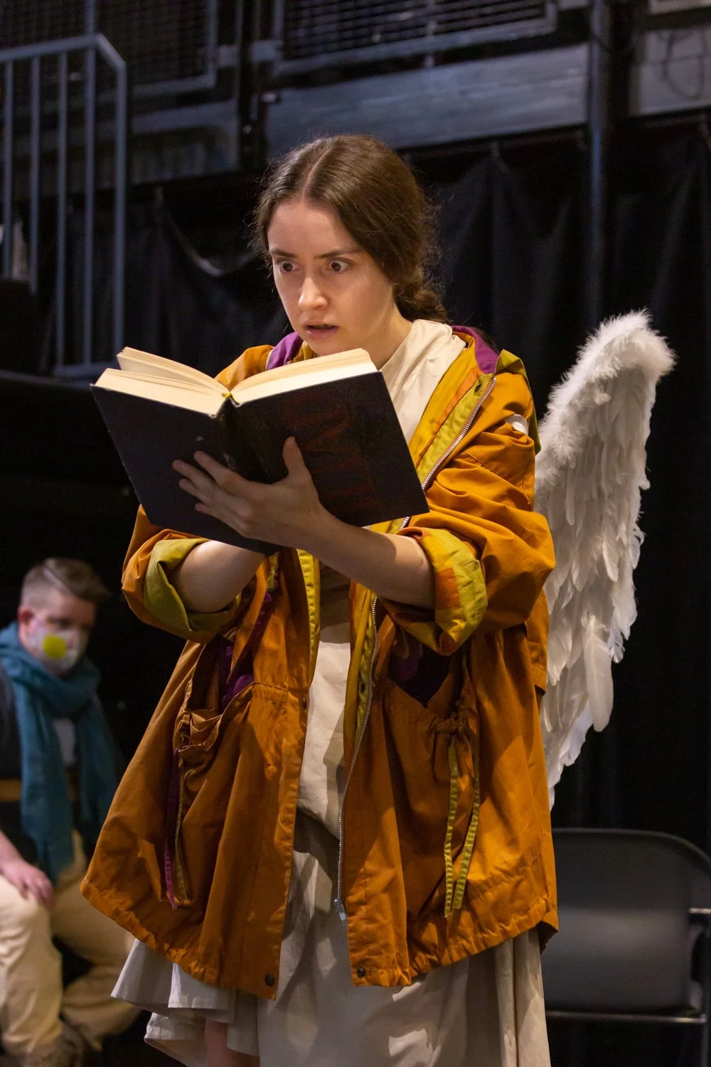 A woman with long brown hair wearing a yellow jacket with a purple backpack looks surprised as she reads a book. She has white angel wings attached to her back and is in a dimly lit theater or stage setting. In the background, there is a man sitting 