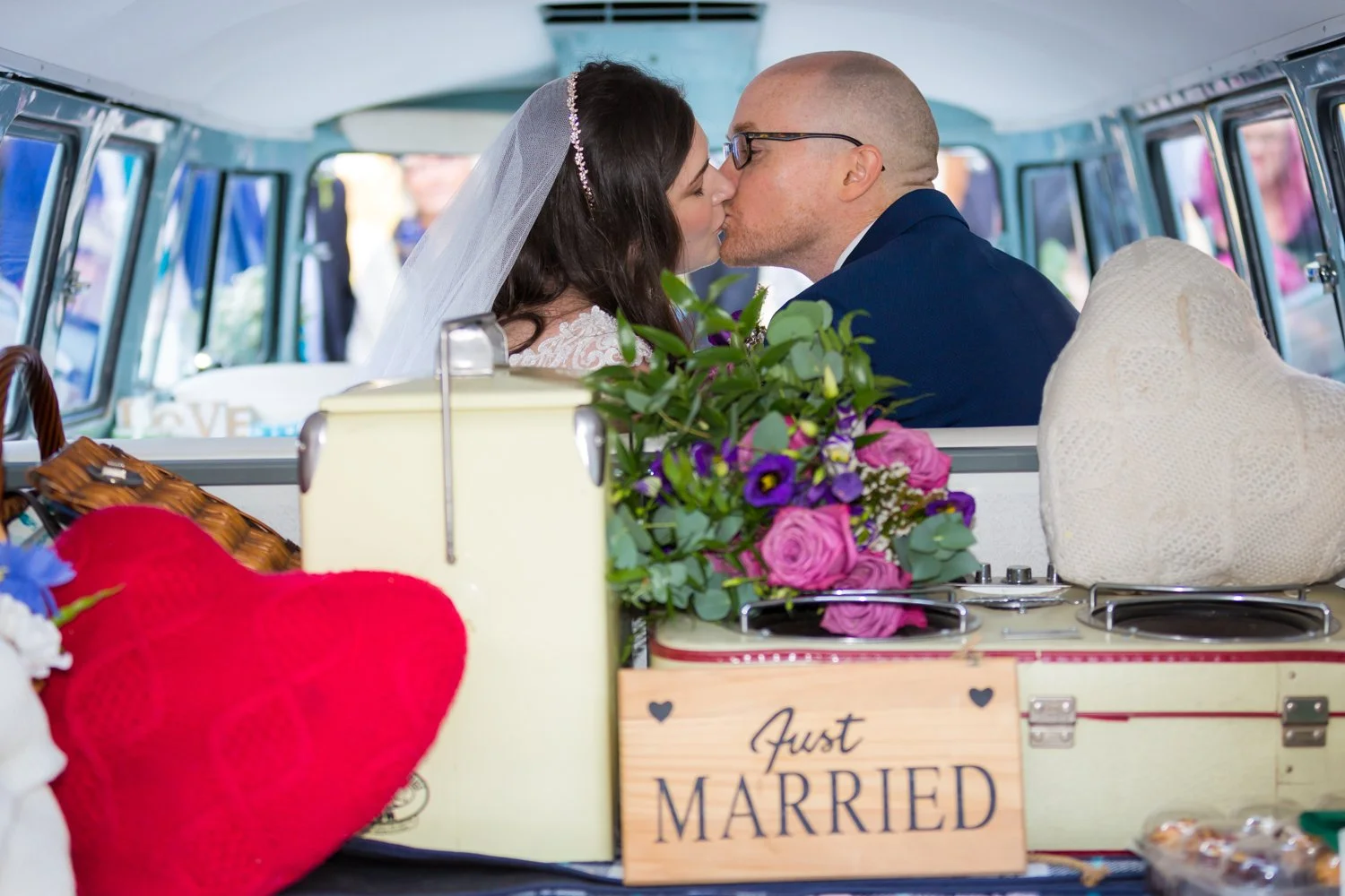 A newly married couple in wedding attire sharing a kiss inside a decorated vehicle, with wedding flowers, a sign that says 'Just Married,' and other wedding-themed items in the foreground.