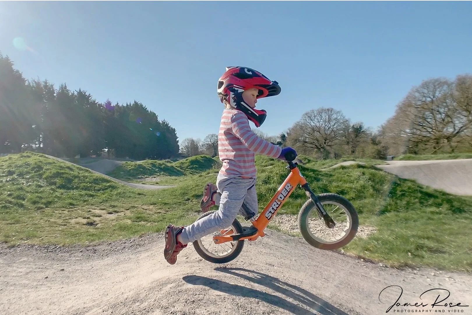 Child wearing a helmet and riding a balance bike on a dirt track in a park on a sunny day.