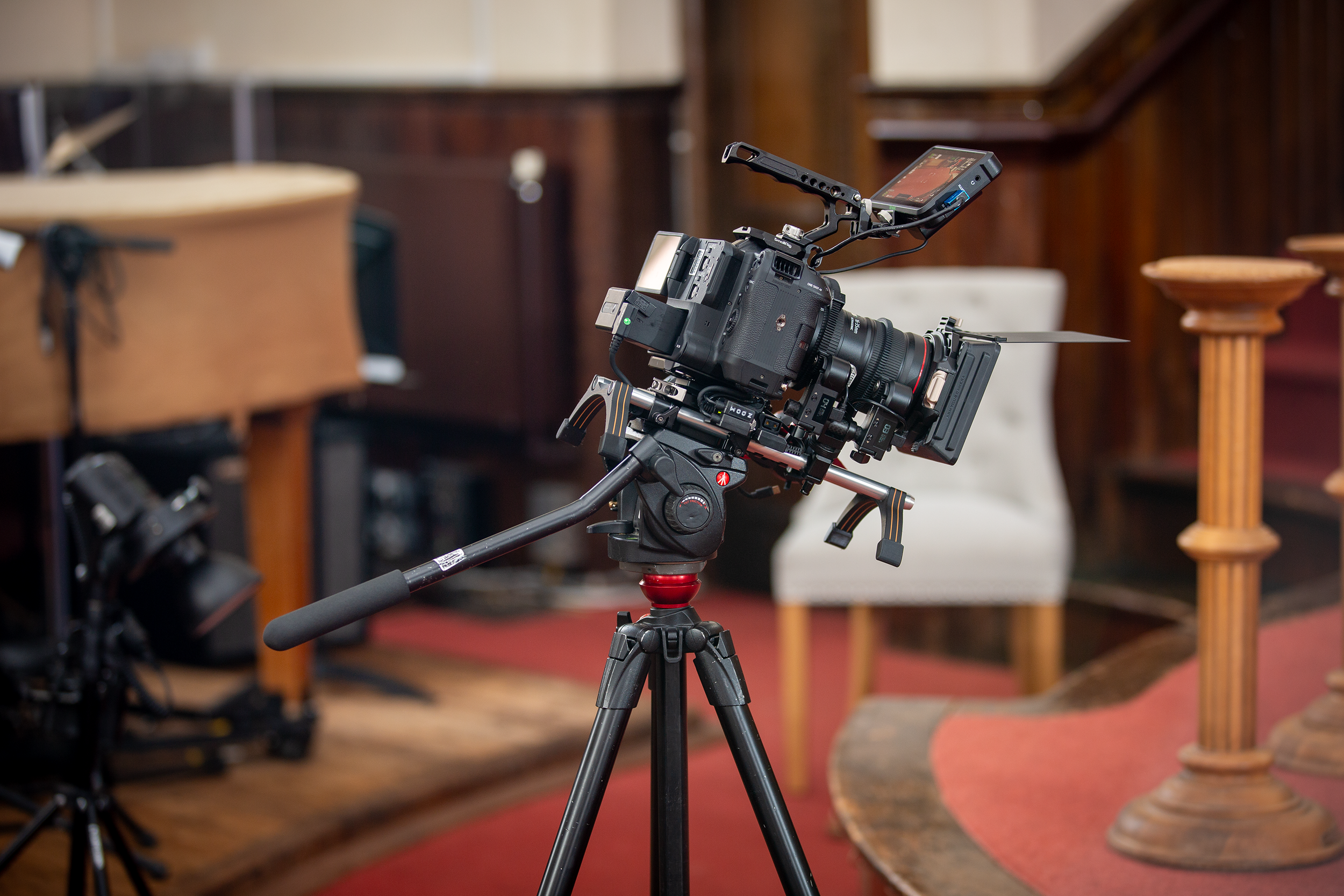 Professional video camera on a tripod set up indoors, with a blurred background of furniture and walls.