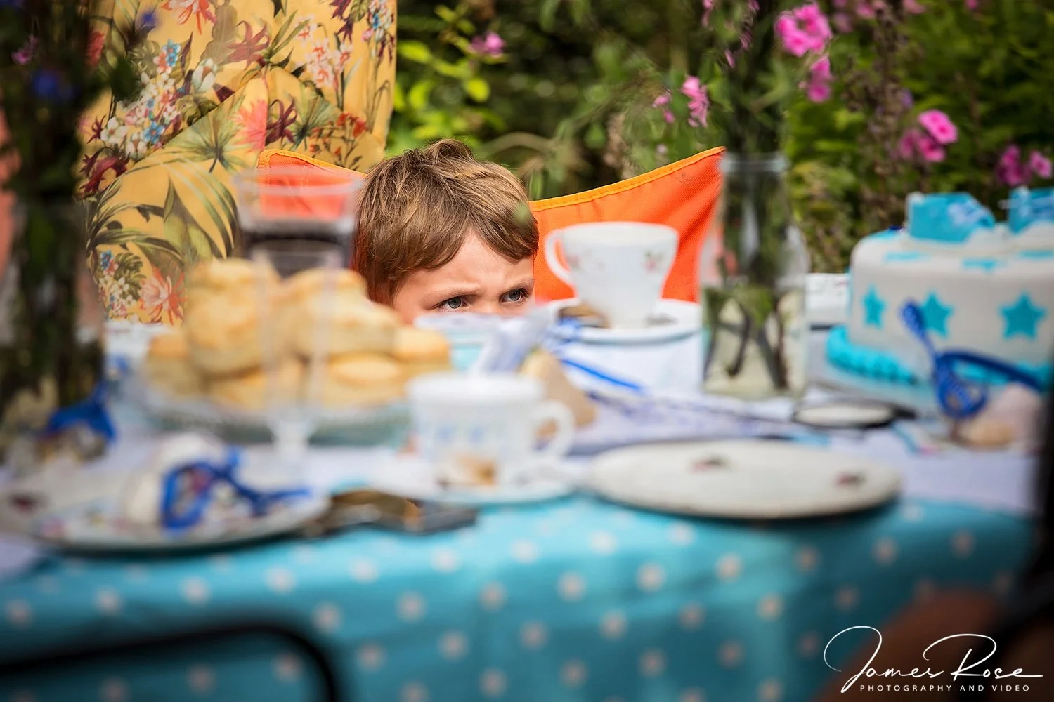 A young boy peeking over a table set for a tea party outdoors, surrounded by teacups, plates, snacks, and decorations, with a garden background.