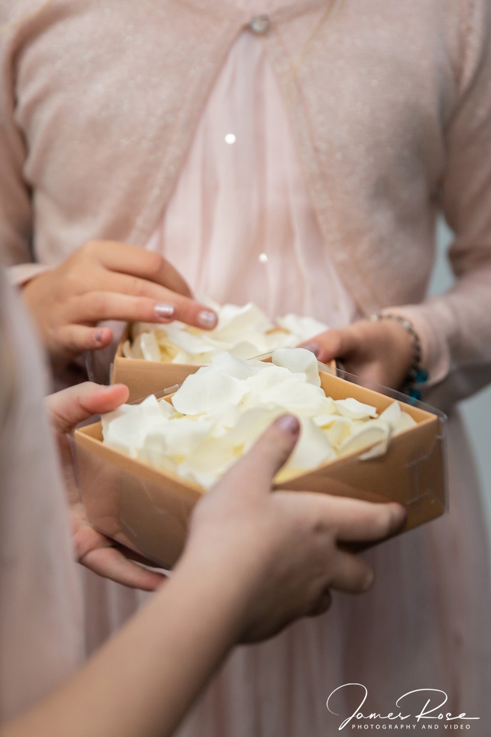 Close-up of hands holding a box of white flower petals, with a person in a pink dress or cardigan in the background.