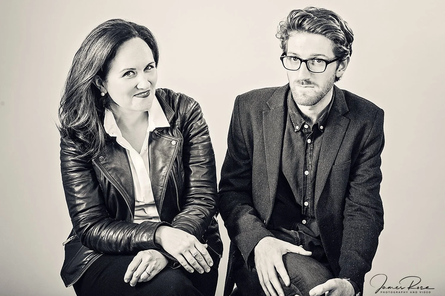 A black-and-white photo of a woman and a man sitting next to each other, both dressed smartly. The woman is wearing a leather jacket over a white shirt, with dark lipstick and pearl earrings. The man is wearing glasses and a dark blazer over a button