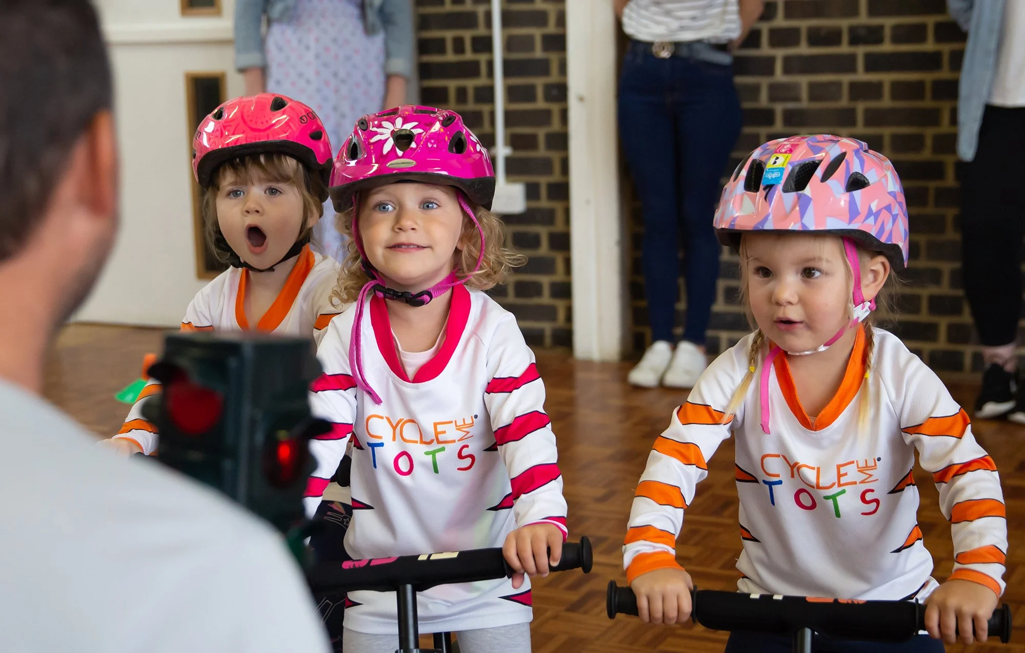 Three young children wearing helmets and cycling jerseys, sitting on balance bikes indoors, participating in a cycling activity. An adult appears to be instructing or talking to them.