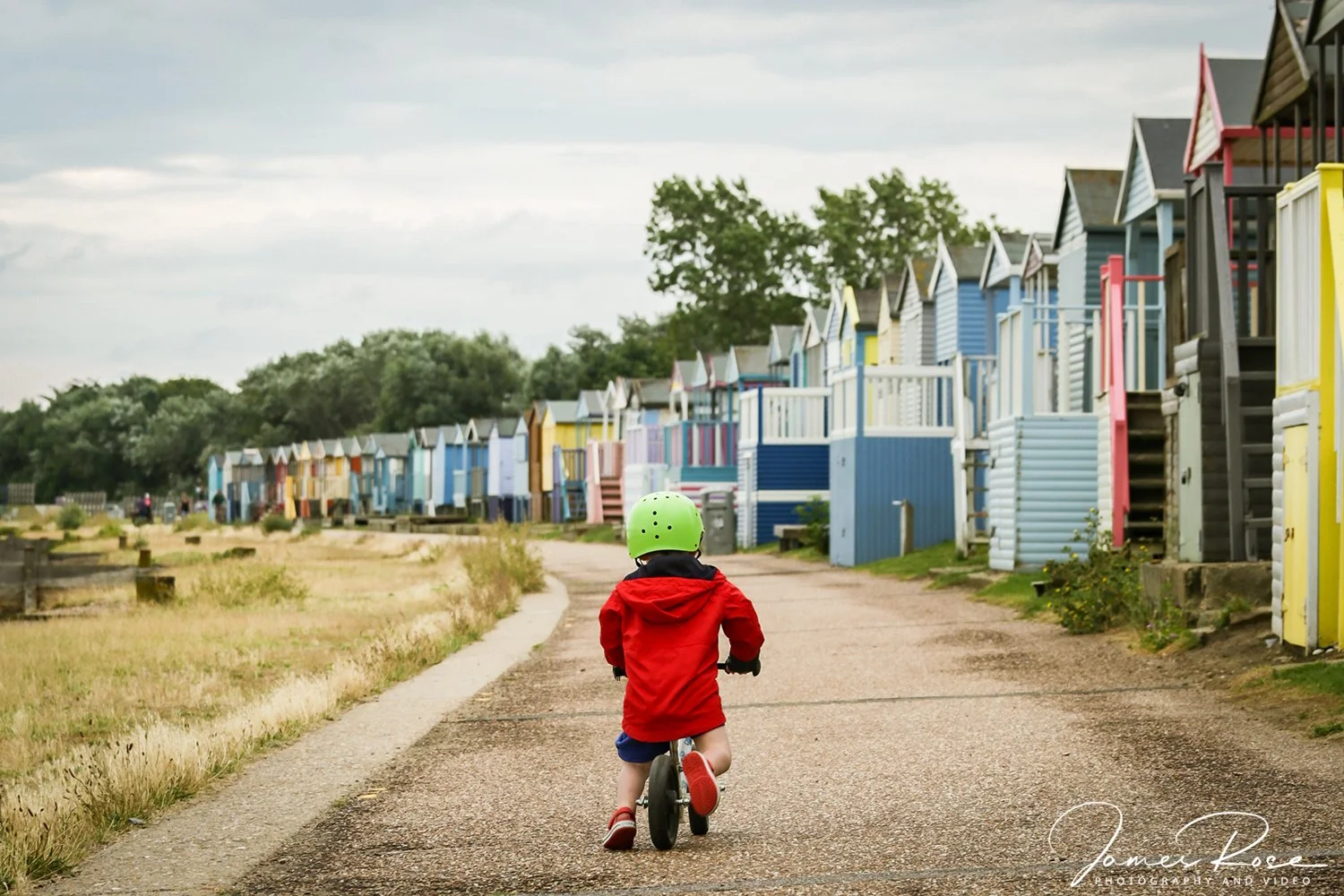 A young child wearing a green helmet and red jacket riding a balance bike on a paved path, with colorful beach huts along the side and a cloudy sky above.