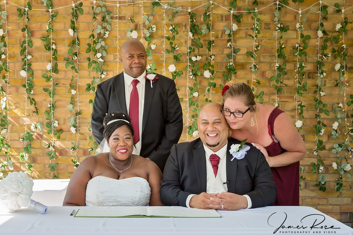 A group of five people at a wedding, with a bride and groom sitting at a table, tilting her head towards him. Two women and one man stand behind them. They are in front of a brick wall decorated with green leaves and white flowers.