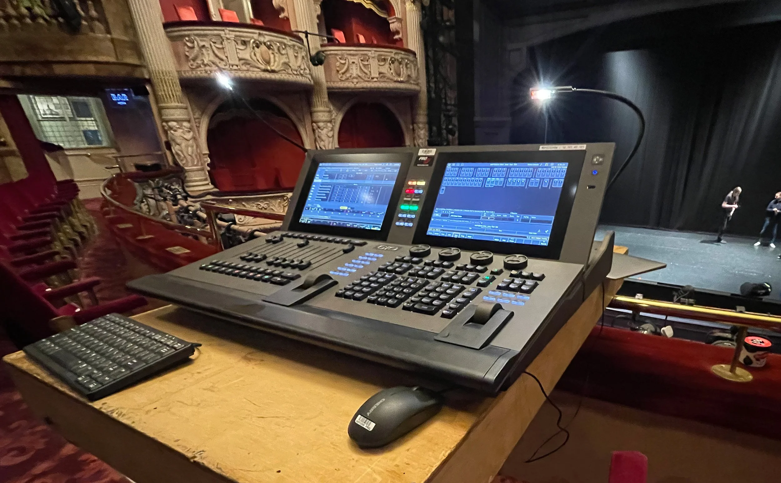 The image shows a stage with two people standing on it, one holding a microphone. In the foreground, there is a lighting and sound control console with screens, buttons, and dials placed on a wooden table. The theater has ornate seats and balcony wit