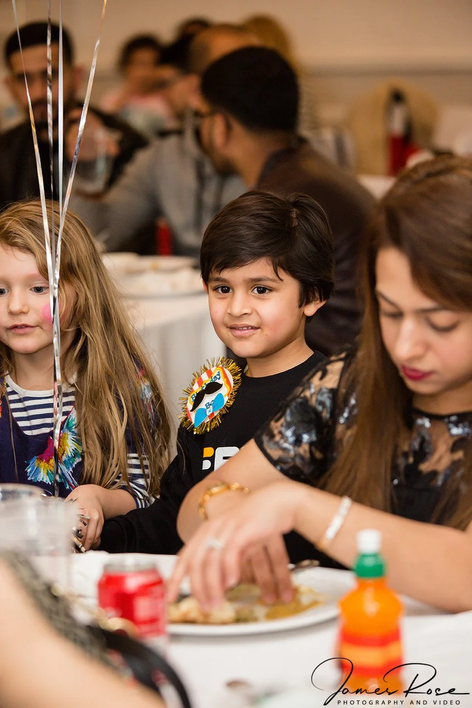Children and adults sit at a table during a celebration. A little boy with dark hair and a necklace featuring a swan is looking ahead, while a girl with long blonde hair and a striped shirt is on his left. In the background, people are talking, and t