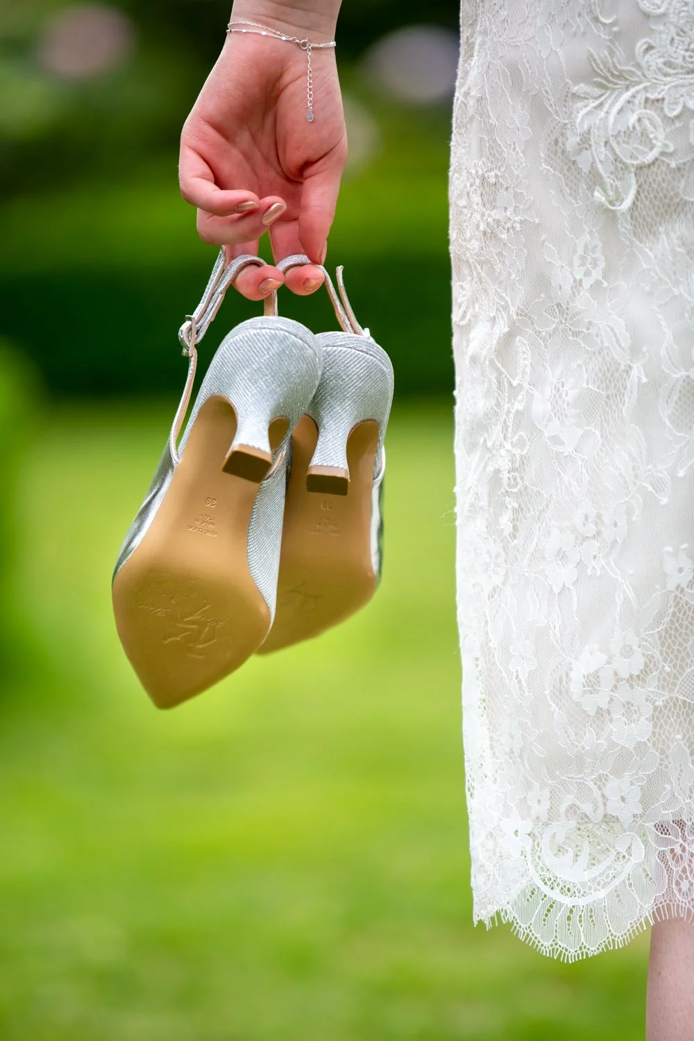 A woman in a lace dress is holding a pair of silver high-heeled shoes by the heels, with a blurred outdoor background.