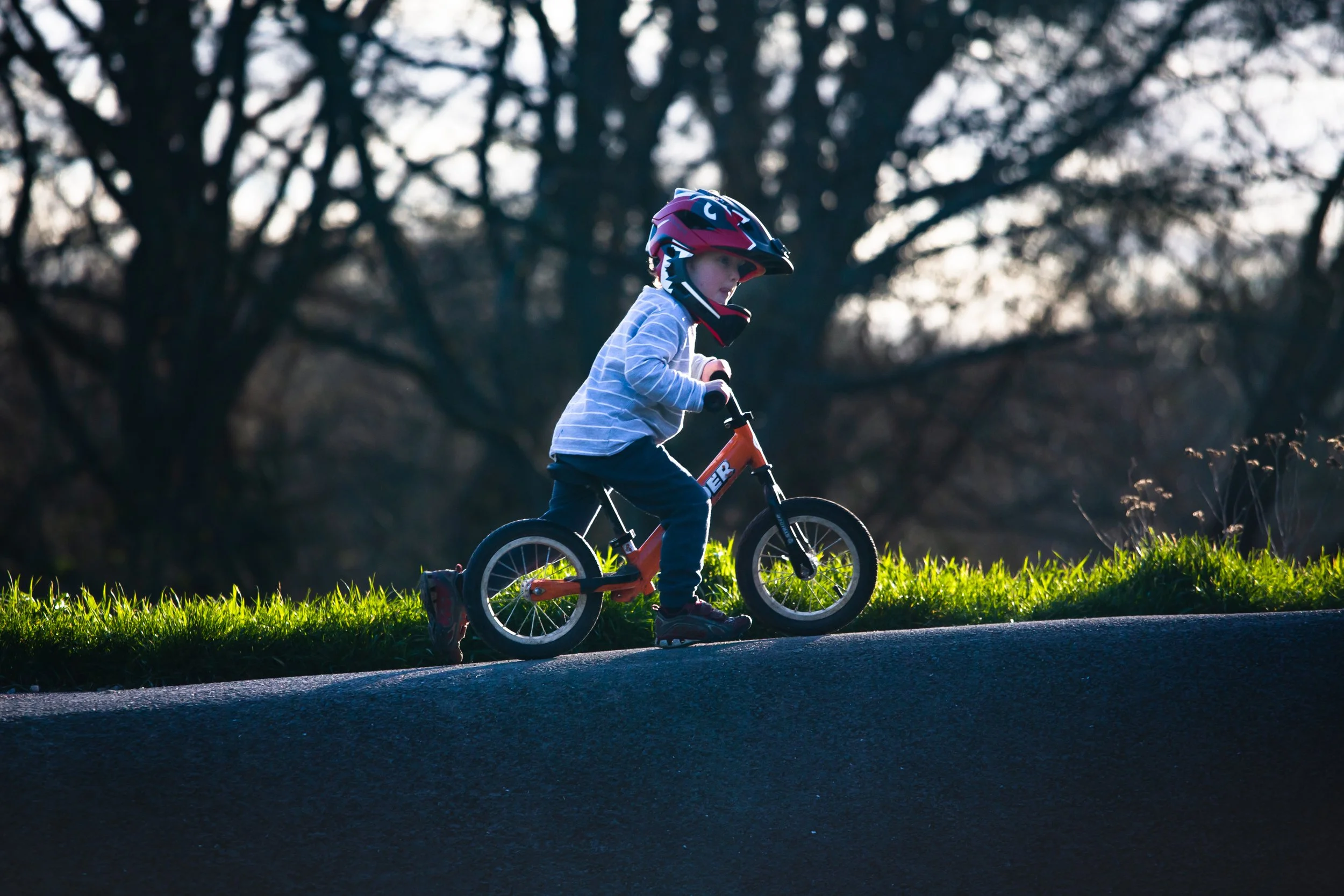 A young child wearing a helmet is riding a small orange bicycle on a path with grass and trees in the background.