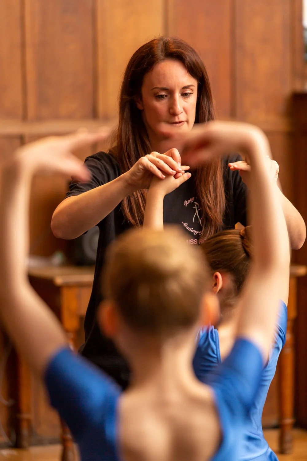 A woman instructing a group of young girls during a ballet class, with the girls stretching their arms overhead.