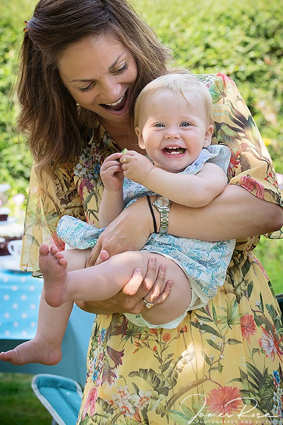 A woman holding a smiling toddler outdoors on a sunny day.