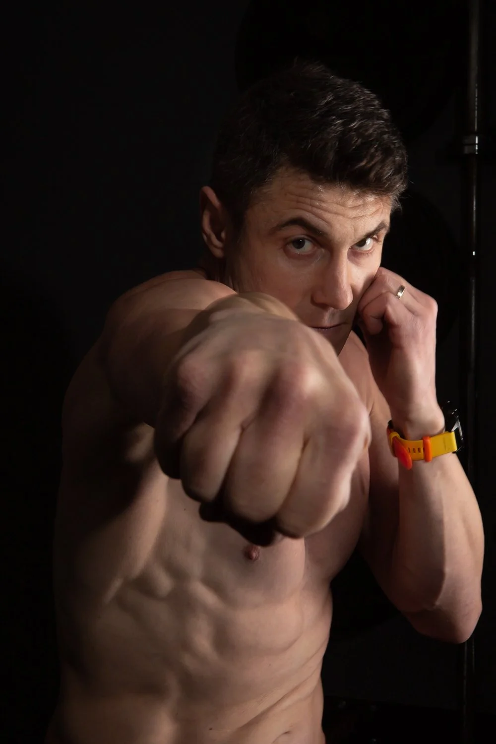 A shirtless man with short dark hair, wearing a yellow watch, is posed with a fist extended toward the camera and his other hand near his face in a boxing stance against a dark background.