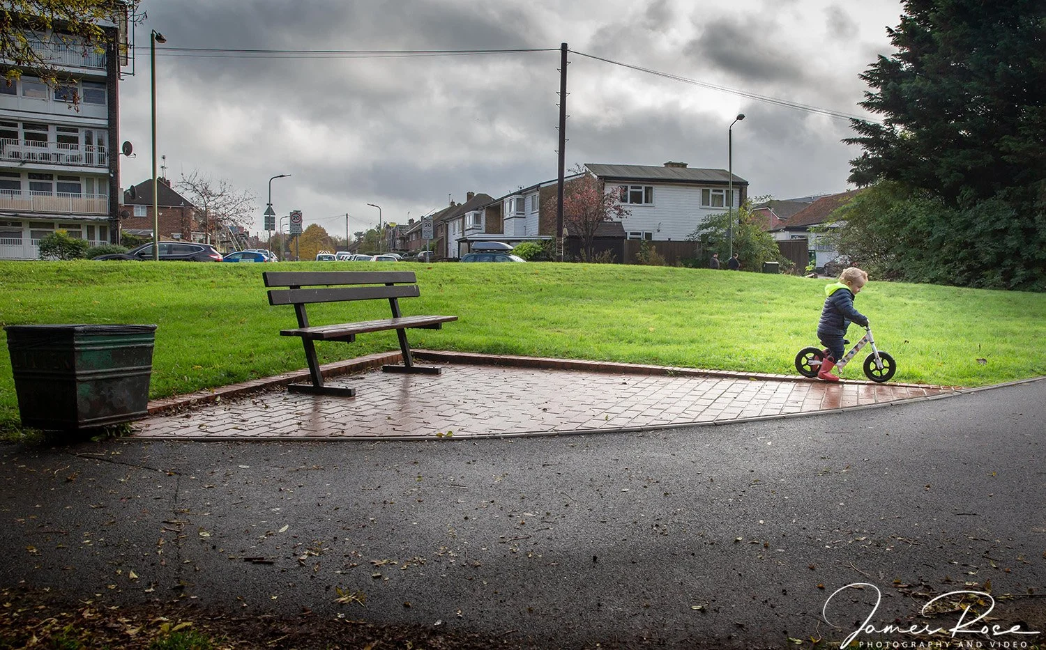A young child riding a balance bike on a paved sidewalk next to a grassy park area with a bench and a trash bin, under a cloudy sky.