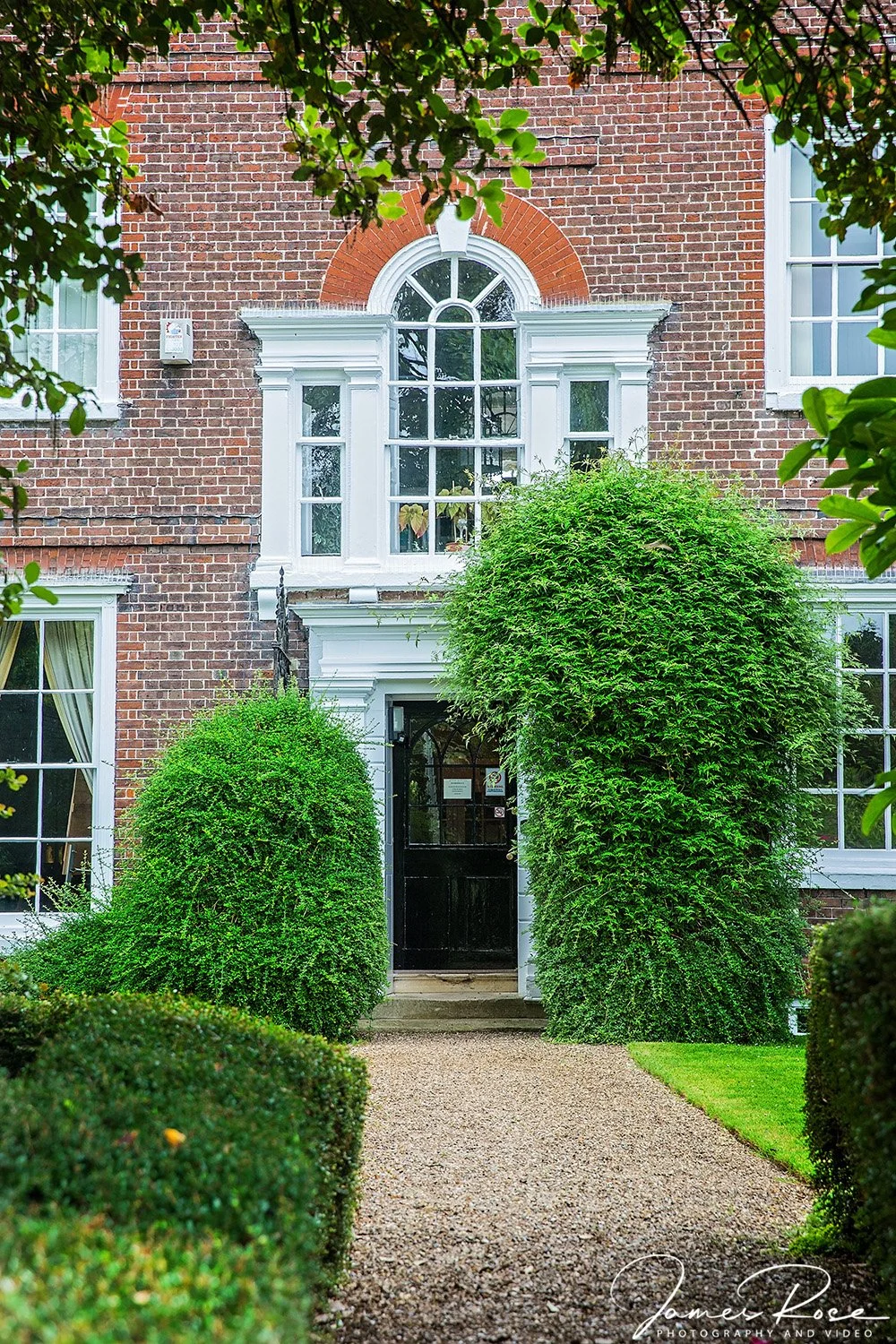 Front view of a brick house with white trim, large window above the door, and two green shrubs flanking the black front door, framed by a pathway and greenery.