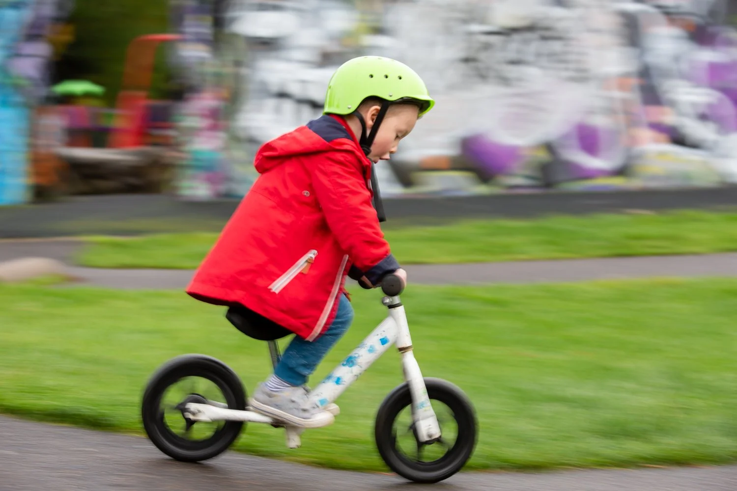 A young child wearing a green helmet and red jacket rides a white balance bike on a paved path with green grass on the side, blurred colorful background.