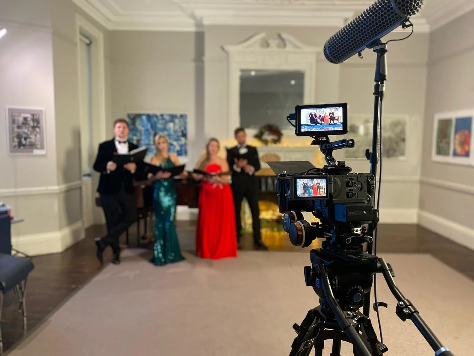 A film crew is filming a group of four singers in formal attire, standing by a fireplace in a decorated room. The camera and microphone are focused on the singers.