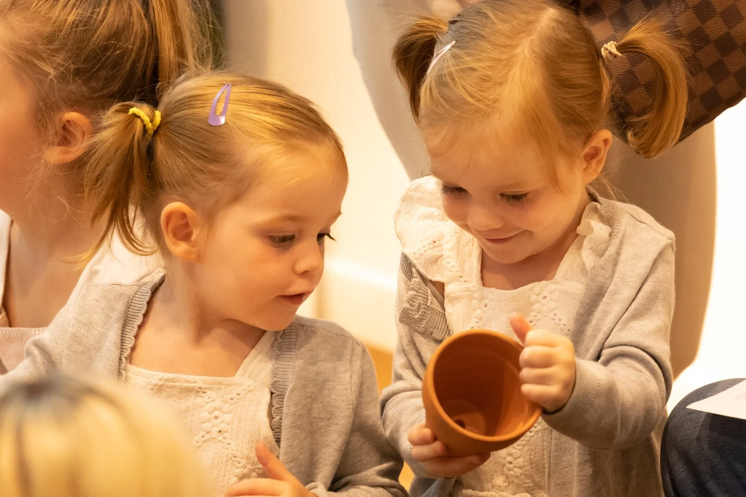 Two young girls with blonde hair and light-colored clothes looking into a small brown cup held by one girl, with an adult partially visible nearby.