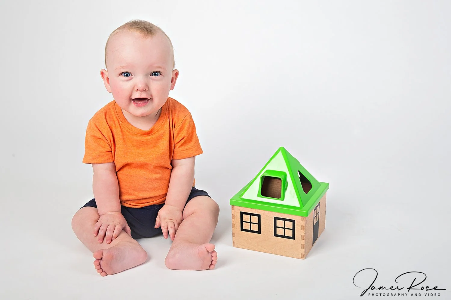 A smiling toddler wearing an orange shirt and navy shorts sitting on a white background next to a wooden toy house with a green roof