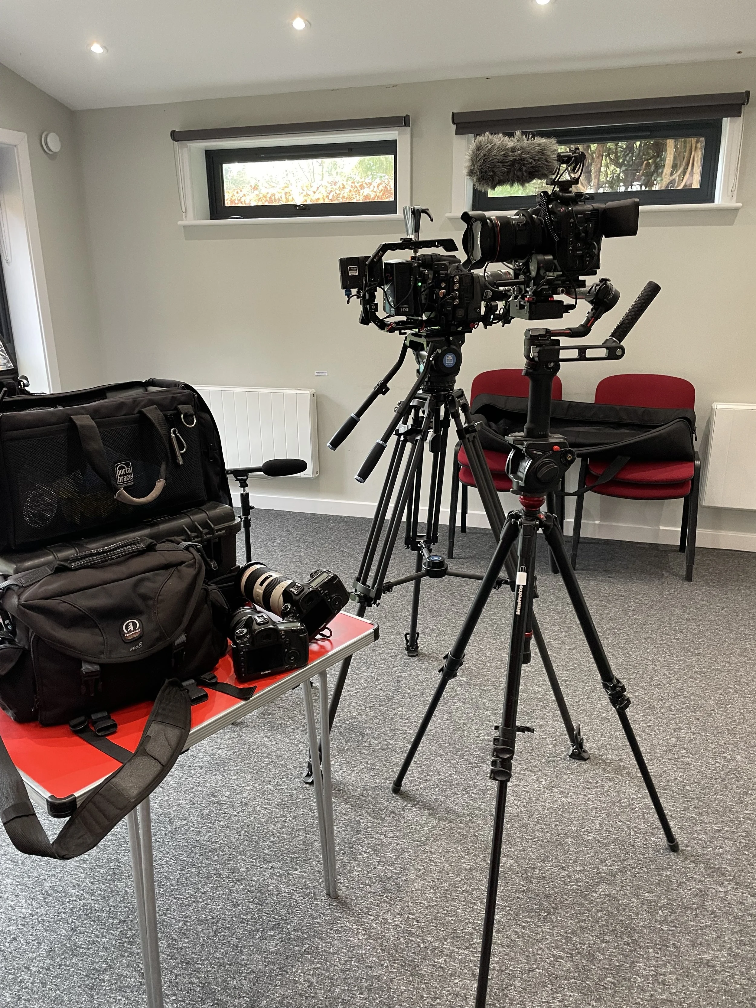 Camera equipment set up in a room with gray carpet, white walls, two small rectangular windows, a black camera mounted on a tripod, and a table with a backpack, a camera, and a camera lens.