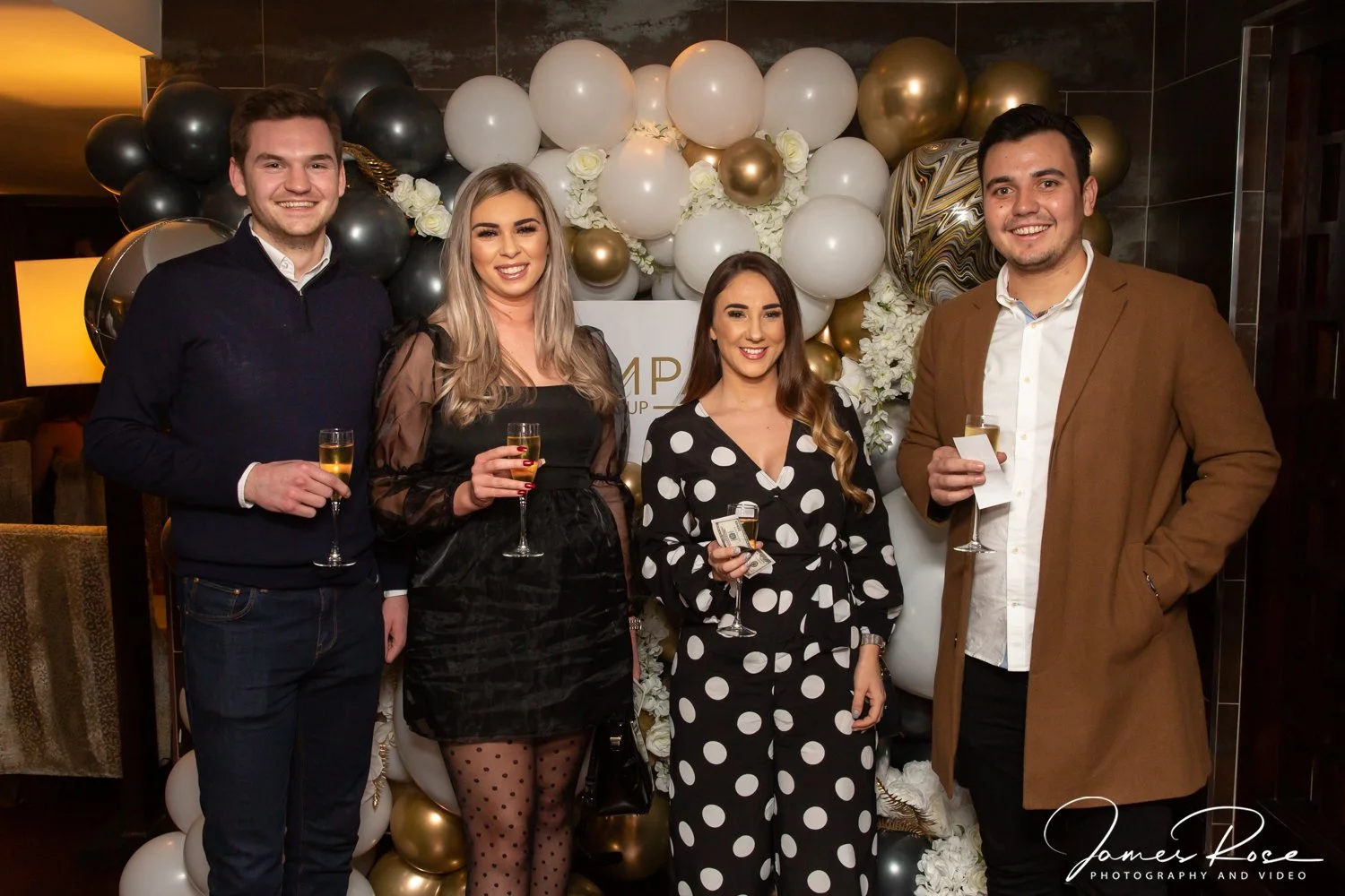 Four people standing in front of a backdrop made of balloons and white flowers, holding glasses of champagne. Two men and two women, dressed in semi-formal attire, smiling at the camera at a celebration event.