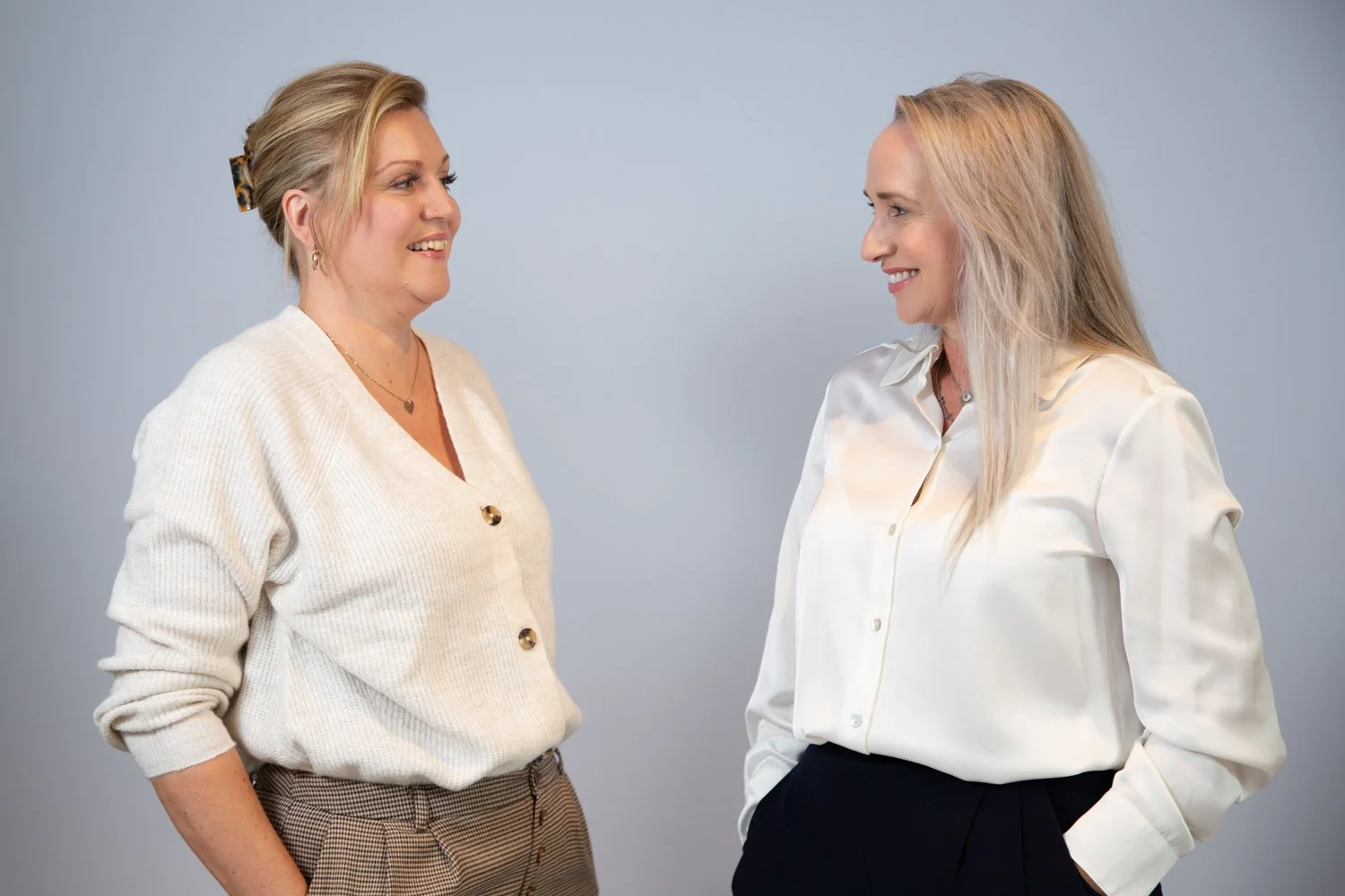 Two women smiling and facing each other in conversation, standing against a plain light gray background.