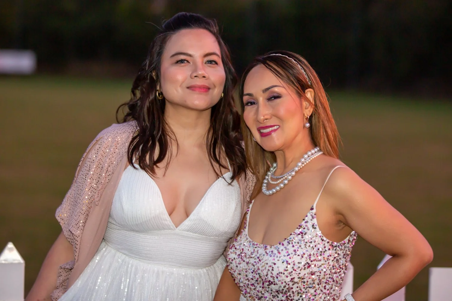Two women standing close together outdoors during evening, smiling. One in a white dress with sheer sleeves, the other in a floral dress with pearl necklaces and earrings.