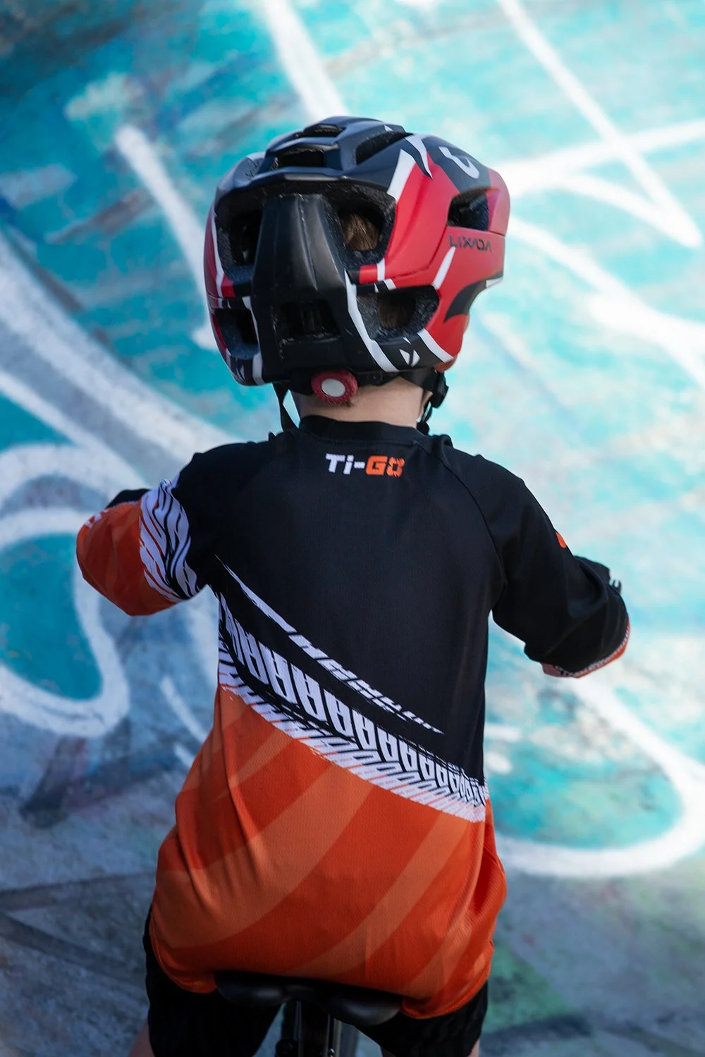 Child wearing a black, orange, and white jersey and an orange and black shorts, with a black and red helmet, riding a bike in front of a colorful graffiti wall.