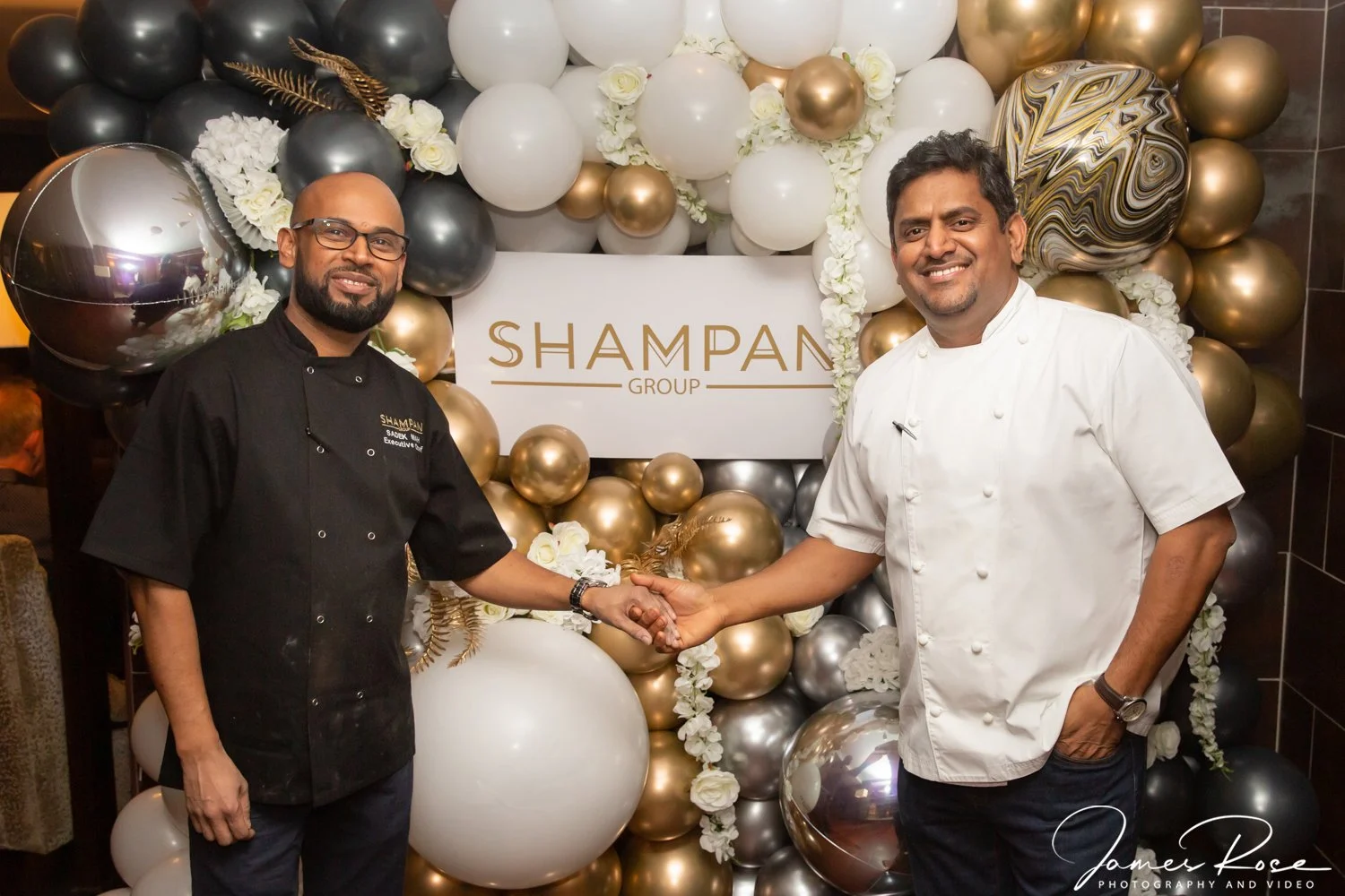 Two men in chef uniforms shaking hands in front of a balloon decoration and a sign that reads "Shampan Group" at a celebration event.