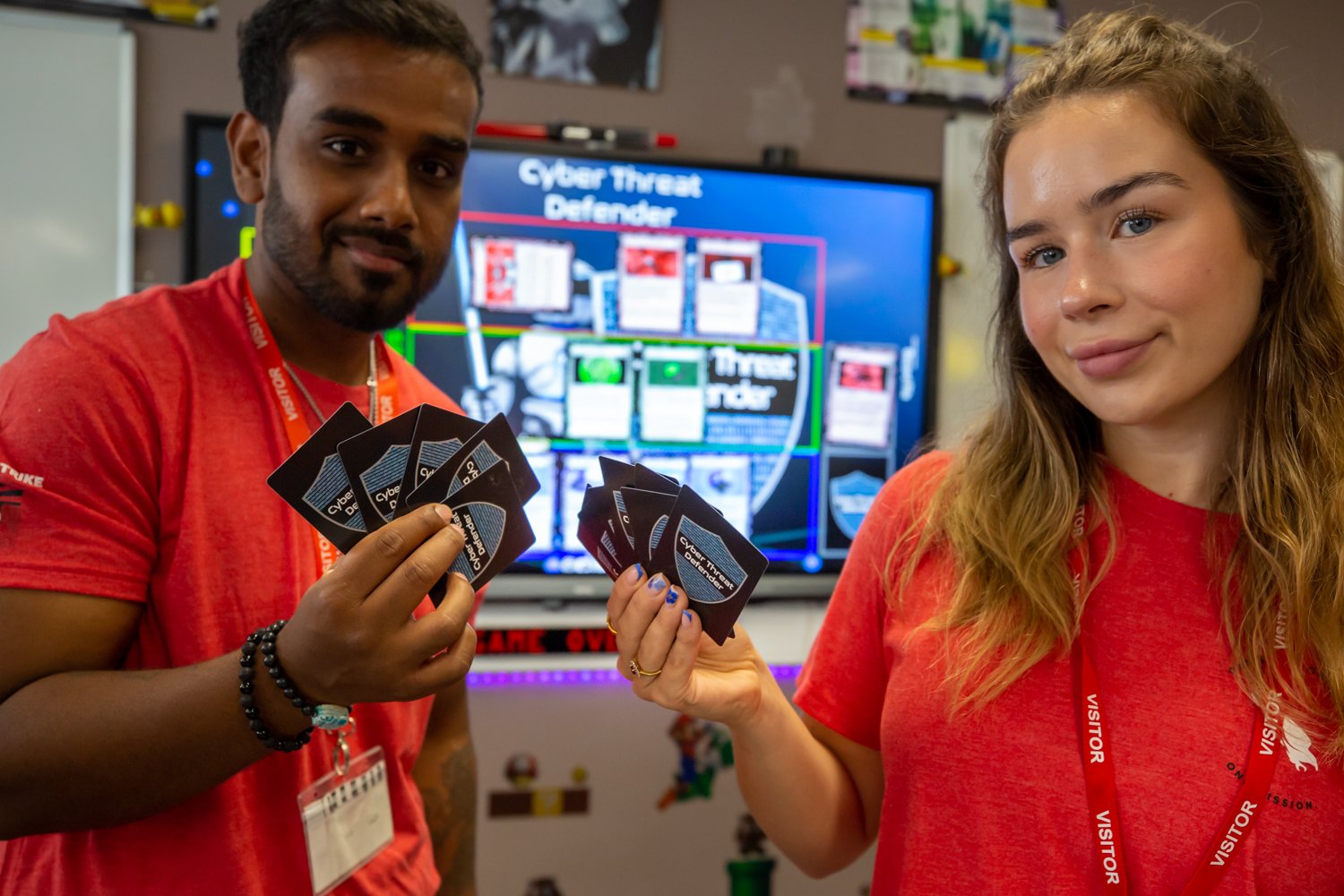Two individuals holding playing cards at a cybersecurity event with a digital screen displaying 'Cyber Threat Defender' in the background.