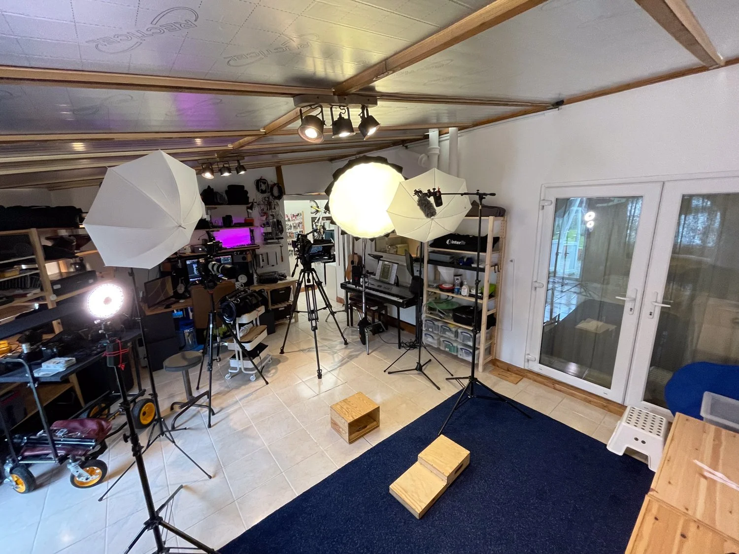 A home studio setup with lighting equipment, cameras on tripods, a piano, shelving with electronic gear, and a blue rug in front of white sliding doors.