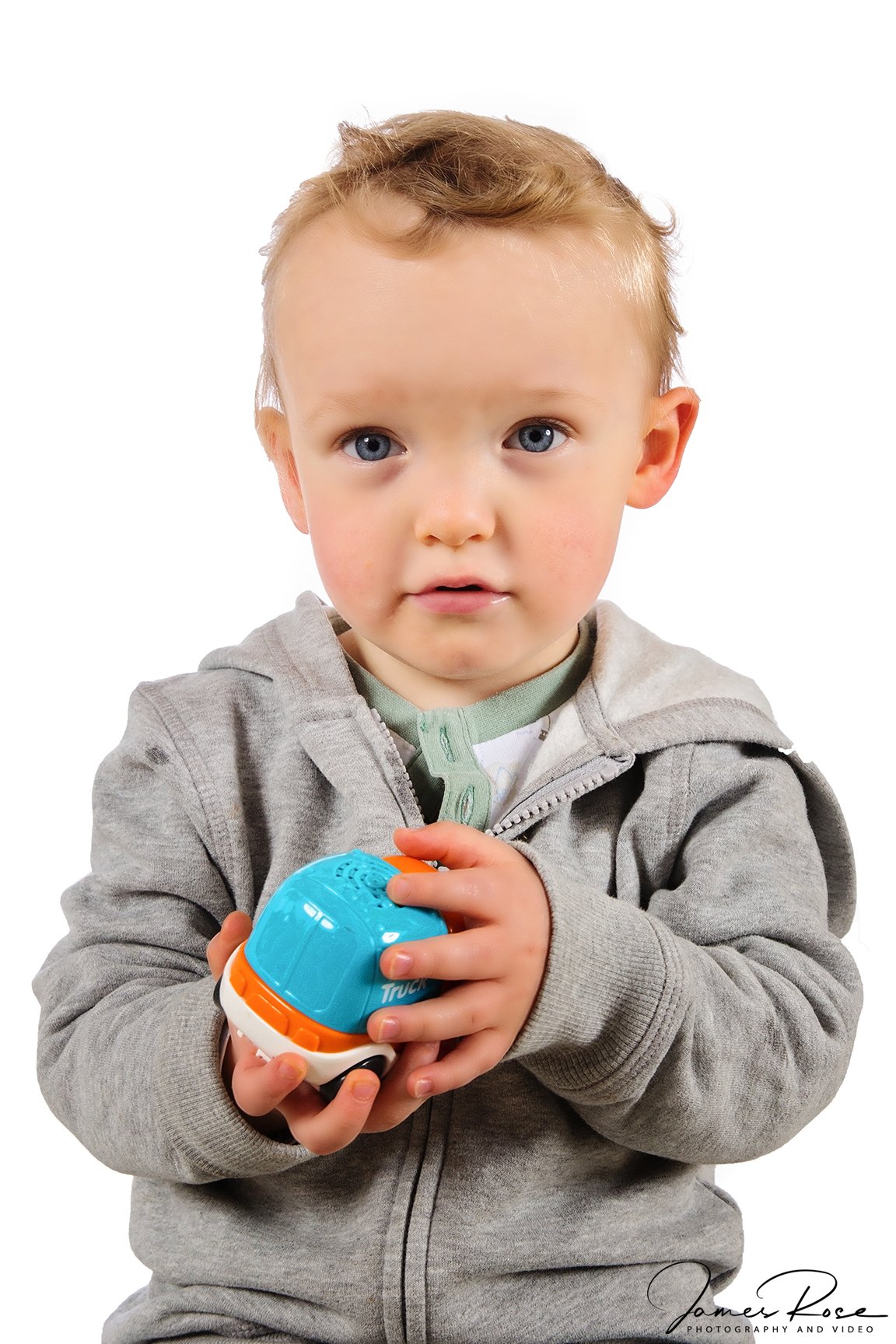 A young boy with blonde hair and blue eyes wearing a gray zip-up hoodie, holding a small blue and orange toy.