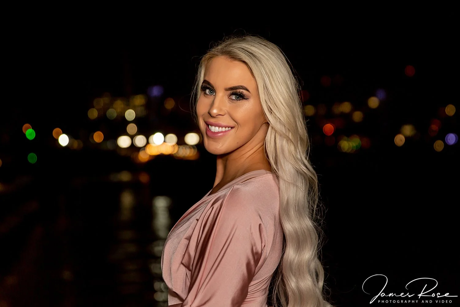 A smiling woman with long blonde wavy hair wearing a pink satin dress standing outdoors at night with city lights blurred in the background.