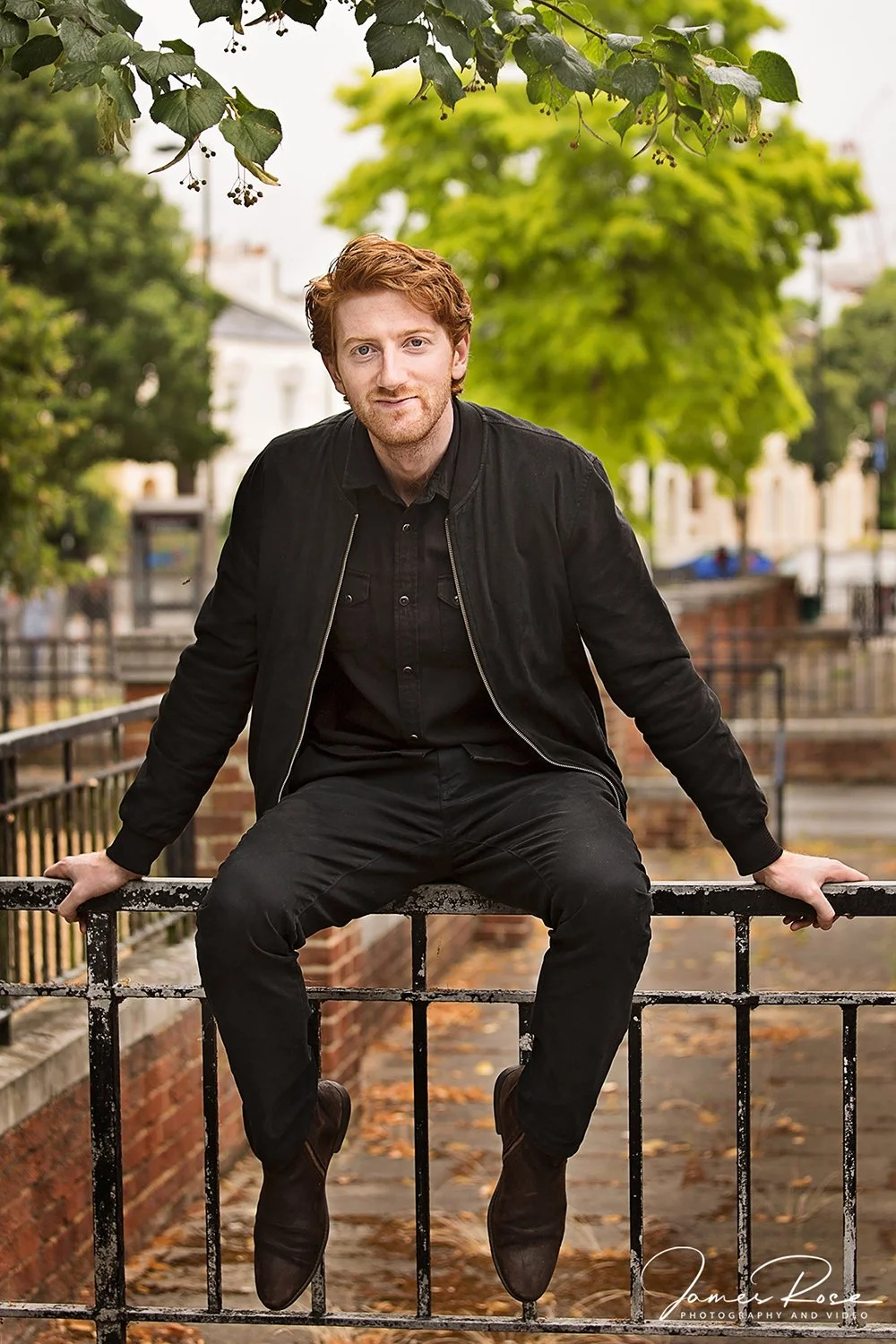A young man with red hair and a beard, wearing a black jacket and black pants, sitting on a black railing outdoors with green trees and buildings in the background.
