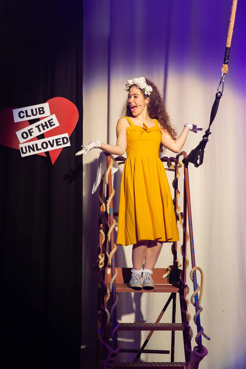 A woman on stage wearing a yellow dress, white gloves, and a flower crown, standing on a podium with a harness and ropes, with a sign that reads 'Club of the Unloved' and a broken red heart in the background.