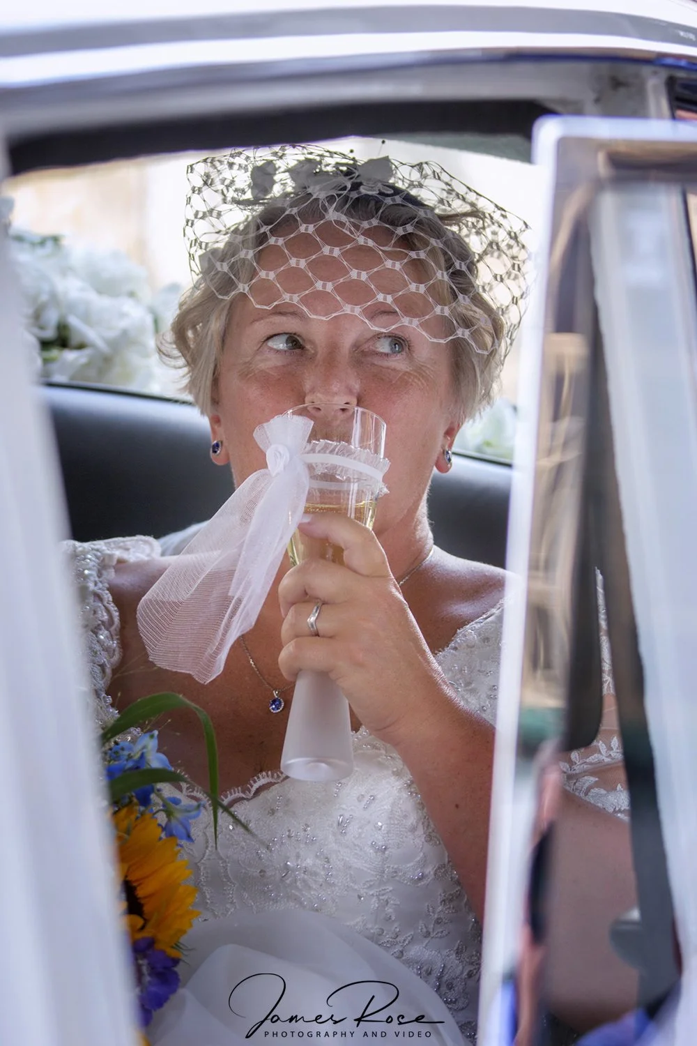 An older woman in a wedding dress and veil sits inside a car, drinking champagne from a flute with a decorative ribbon, surrounded by flowers.