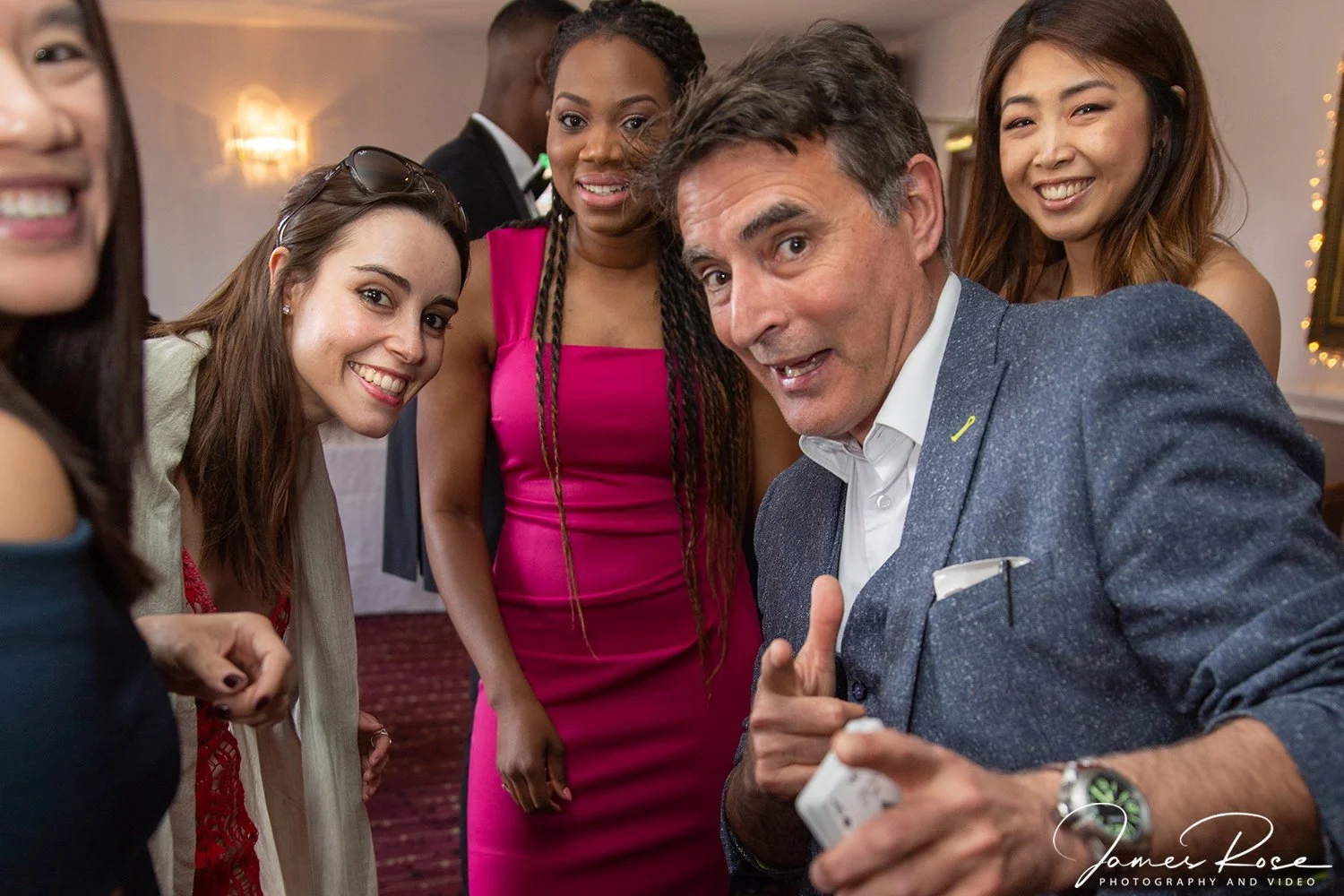 A group of five adults, four women and one man, smiling and gathered closely together in a social setting, with the man showing something on a small device, possibly a phone or a card, to the women.