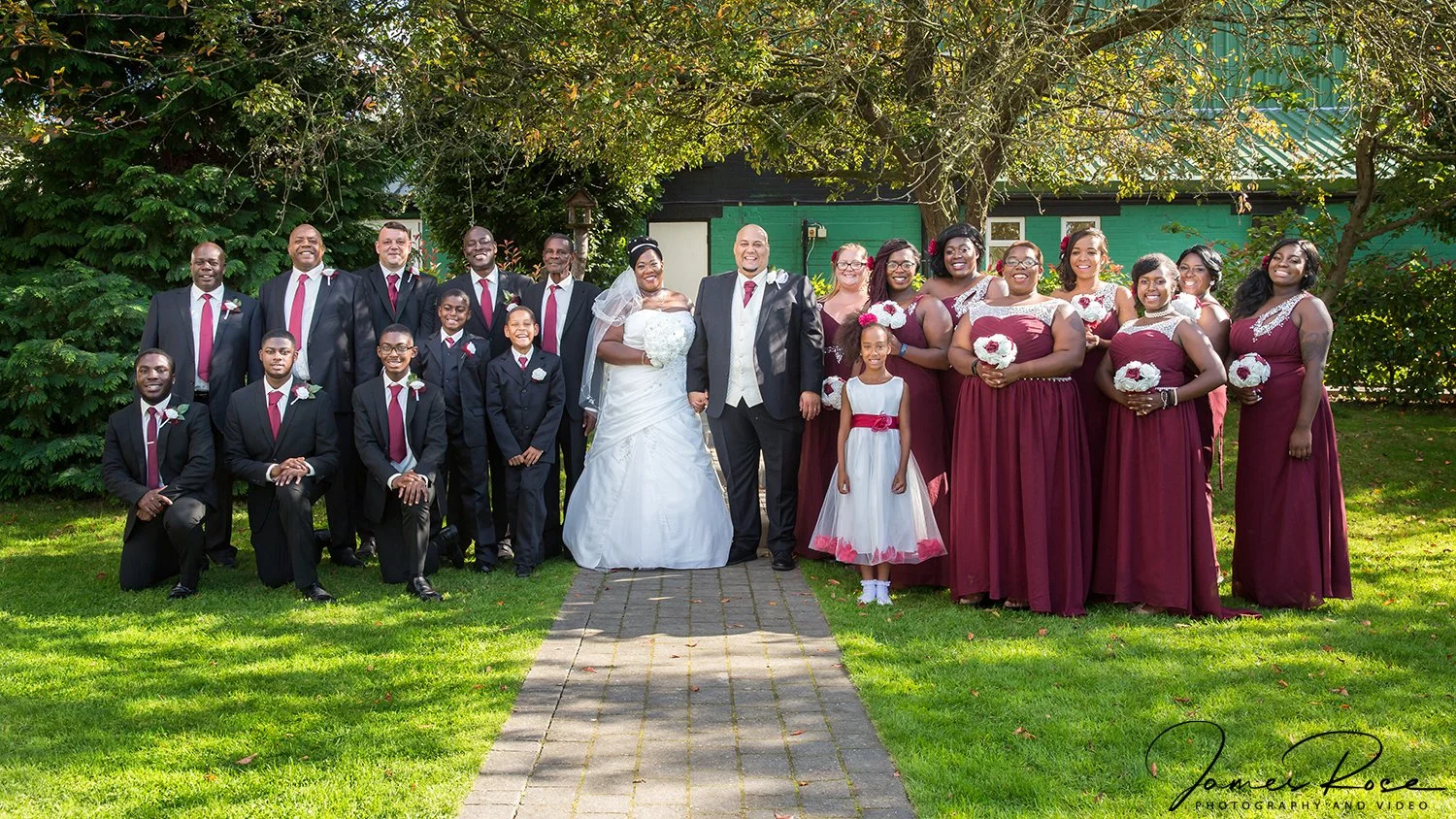 A wedding party outdoors with the bride and groom at the center, surrounded by family and friends. The bride is wearing a white wedding dress and the groom is in a black suit with a red tie. The bridesmaids are dressed in burgundy gown with floral ac